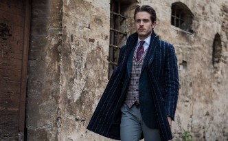 The ice colored trousers by Yan Simmon match the gray and burgundy checkered waist coat, presented here with a blue herringbone jacket. The blue coat by Hevò is made of wool and pinstriped fabric